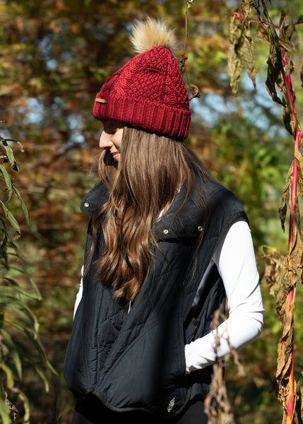 Woman wearing Eco Spurr Beanie in Burgundy with Faux Fur pom-pom, standing outdoors in nature.